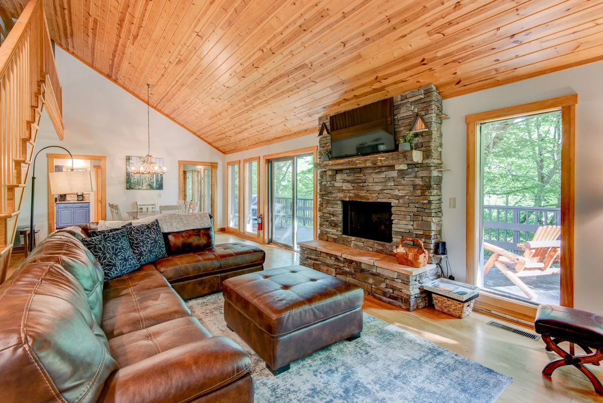 This image offers a broader vantage point of the great room—showing the stone fireplace with a mounted Smart HDTV, a large sectional sofa for group gatherings, wooden ceiling beams, and sliding glass doors opening to the deck outside. The space is filled with natural light and invites relaxation, entertainment, and connection.