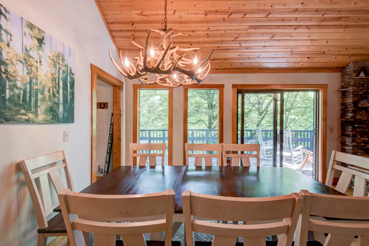 This image displays the spacious dining area with an antler chandelier above a large dining table, surrounded by large windows bringing in natural light. The scene radiates coziness and is ideal for social gatherings, game nights, and family dinners, with visual cues of a seamless transition from living to dining space.