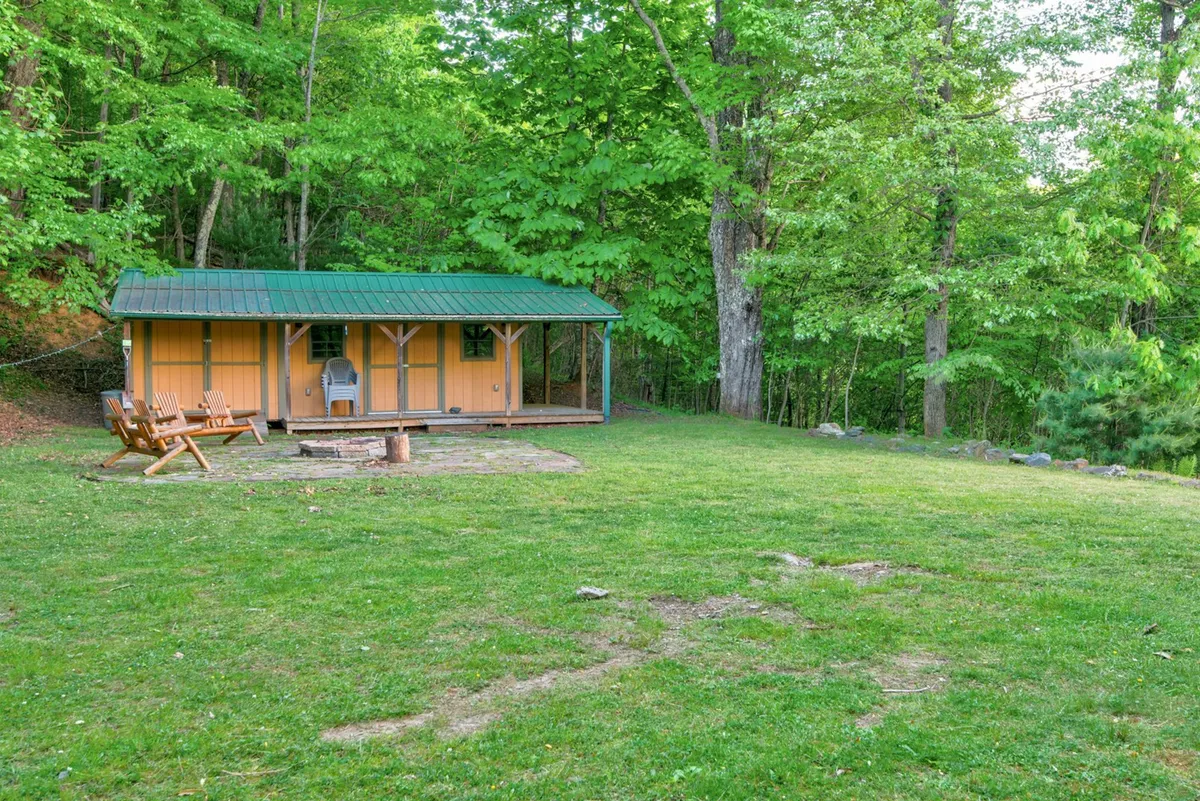 This exterior shot highlights a cheerful yellow cabin with a green roof, a fire pit, wooden chairs, and lush greenery. The open lawn area offers a glimpse of a semi-flat yard, ideal for dogs to play, while the fire pit and surrounding woods suggest evenings spent together outdoors and opportunities for nature walks.