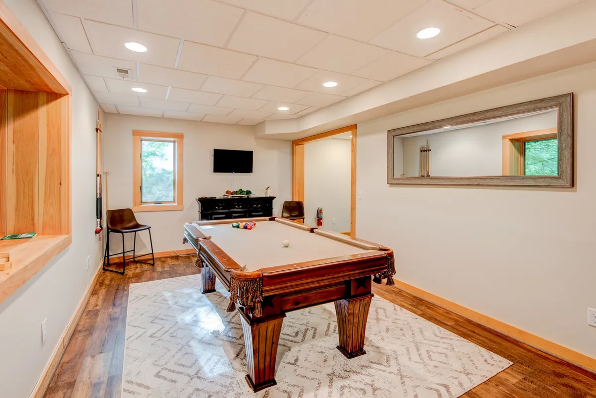A dynamic interior view featuring a pool table, wooden accents, a wall-mounted TV, and a mirrored wall decoration. This space is designed for casual gatherings, offering options for both billiards and movie nights, perfectly matching the idea of a 'secondary living area' for kids, teens, or late-night entertainment.