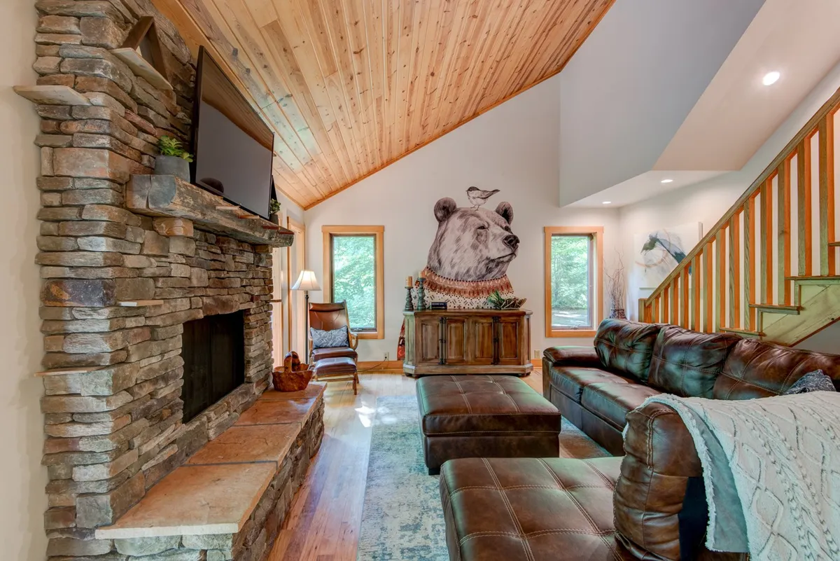 This image showcases the great room's central living area, featuring a dramatic stacked stone fireplace, a prominent bear mural, a comfortable leather sofa, and a wooden coffee table. The space exudes warmth with hardwood floors and artistic mountain-inspired decor, capturing the inviting and communal heart of the home.