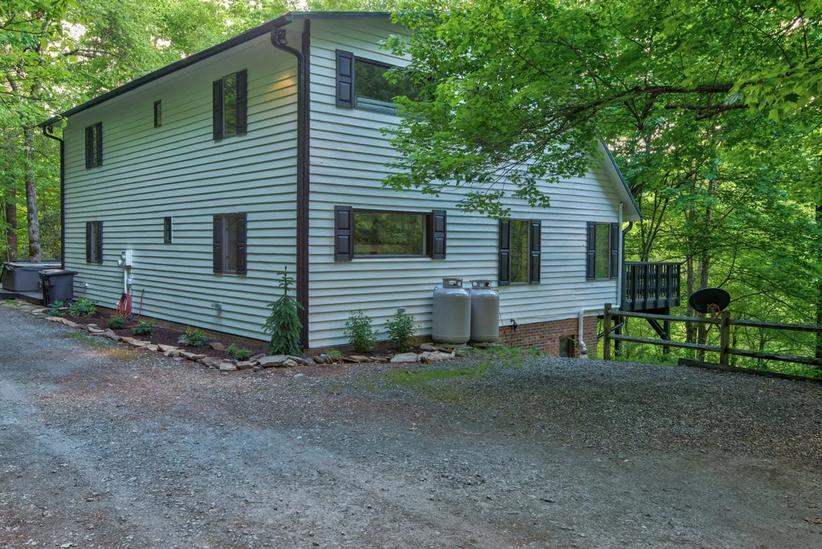The house exterior is shown with large windows and is surrounded by green landscaping and trees, creating a tranquil, nature-filled setting. The porch and garden area highlight the connection to the outdoors.