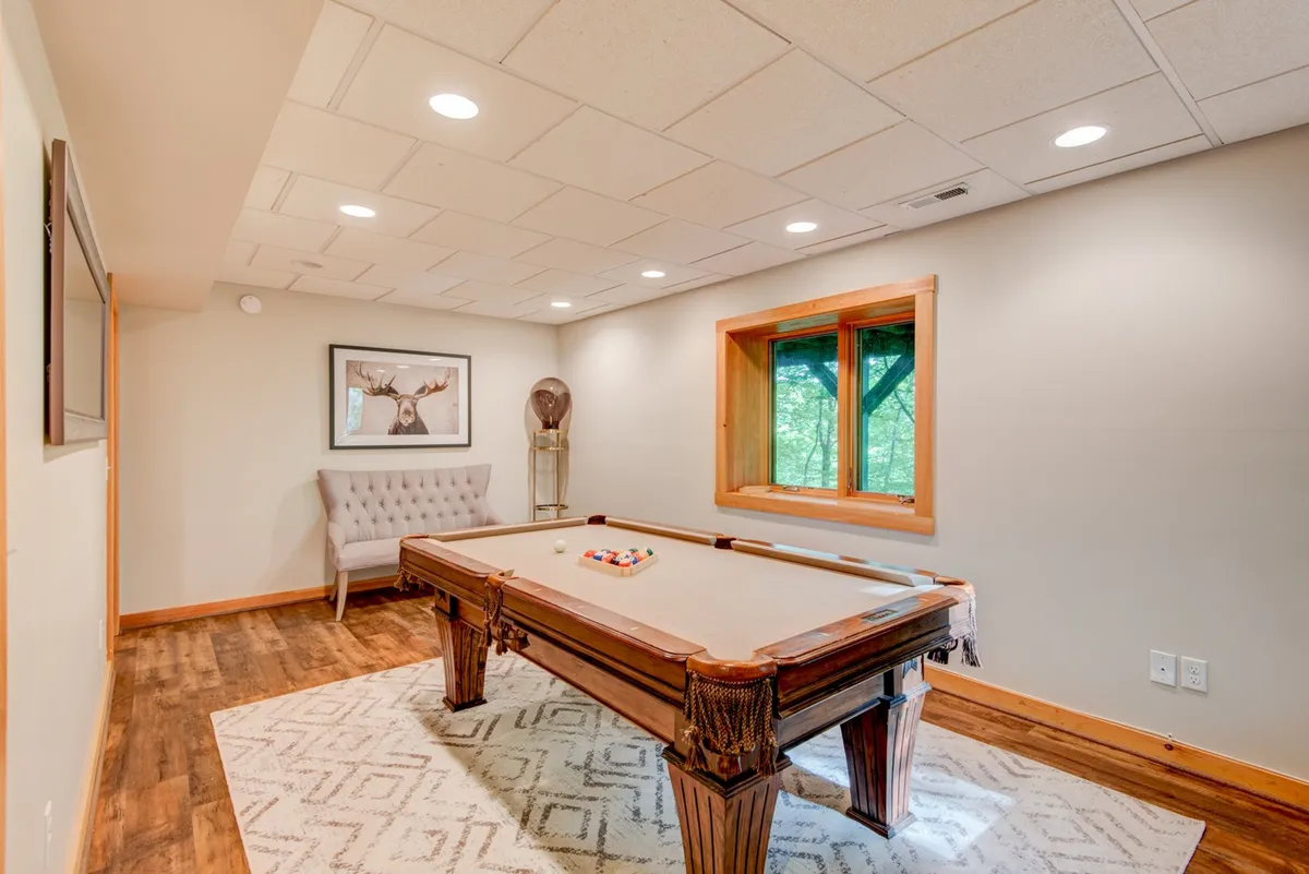 This image captures a vibrant game area with a pool table as the focal point, surrounded by windows letting in natural light and cozy seating for spectators or players to relax. Framed artwork adorns the wall, creating a lively and welcoming atmosphere tailored for group entertainment, casual relaxation, and friendly competition.
