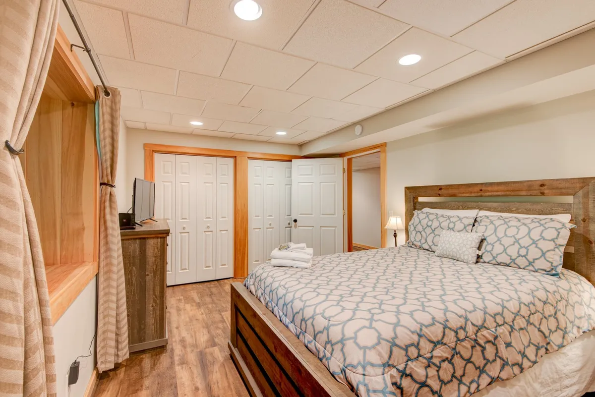 This image showcases a king-sized bed with patterned bedding, complemented by wooden furniture and curtains framing a window. The space exudes tranquility and a sense of comfort, with natural light softening the room's inviting ambiance.