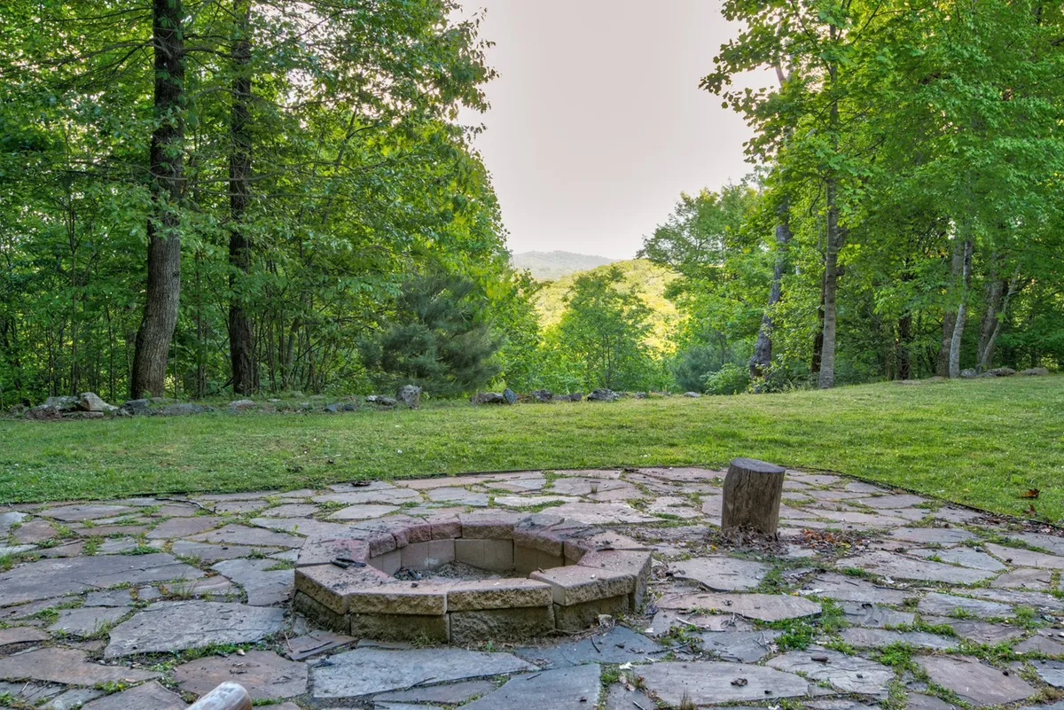 The photo captures a beautiful stone fire pit on a stone patio, encircled by wooden chairs and framed by trees and distant hills. The setting suggests warm gatherings, stargazing, and moments of togetherness, making it ideal for seasonal celebrations.