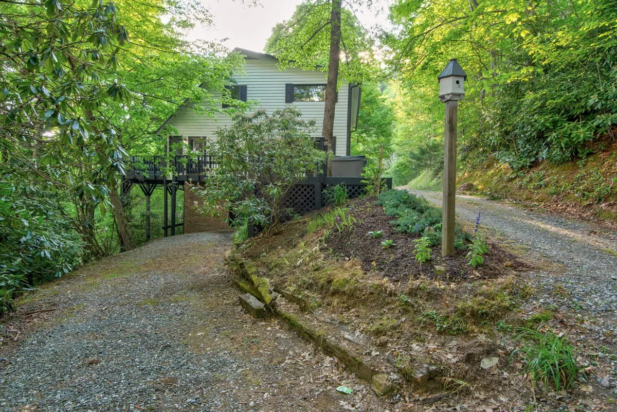This image features the house nestled among mature trees with a gravel driveway and a birdhouse on a post. The lush, green surroundings create a serene, inviting environment that hints at the natural setting and outdoor adventures available for both people and pets. The cozy placement of the house underscores its accessibility to nature and nearby trails.