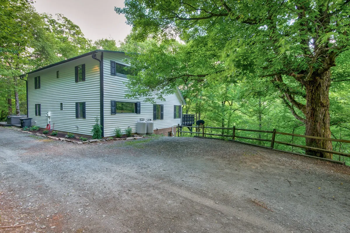 A two-story house surrounded by lush greenery, with a fenced yard, an outdoor seating area, and a gravel driveway. The semi-flat lawn area provides ample space for pets to play while remaining close to the house.
