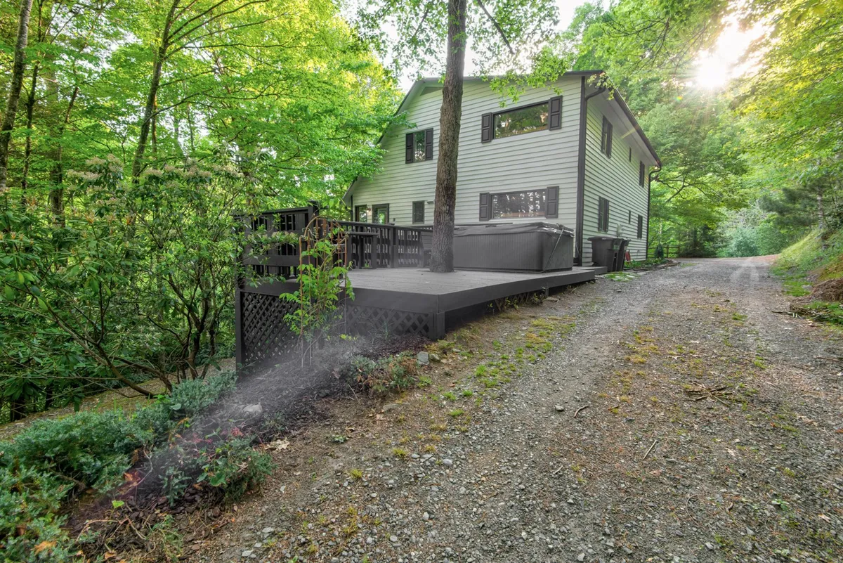 This image showcases a spacious deck nestled among the trees with a hot tub and outdoor seating area. The scene is serene and inviting, highlighting natural surroundings and offering views of the lush forest, perfect for relaxation across all seasons.